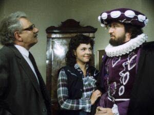 Three people indoors: a suited older man on the left, a smiling woman in a plaid shirt at center, and a bearded man in a purple Elizabethan costume on the right.