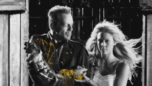 Middle-aged man in a dark suit with yellow paint splatters, looking down at his splattered arm, standing beside a young blonde woman in a dark, wooden barn setting.