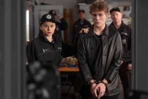 Female police officer with hand on a detainee's shoulder, a man in a black leather jacket handcuffed in the foreground, others visible in the background.