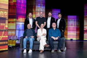 Seven people pose on a brightly colored TV studio set: four men stand behind a sofa while three others sit on the sofa, smiling at the camera.