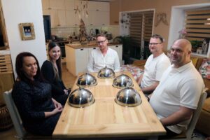 Five adults sit around a wooden dining table in a bright home kitchen, smiling at the camera with cloche covers on the table edgewise in front of them.