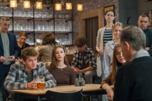 Group of young adults socializing around a table in a busy cafe/bar, with a beer mug in the foreground and shelves of glassware in the background.