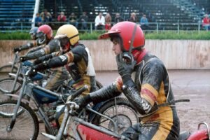 Riders in colorful helmets and muddy gear line up on dirt bikes at a race track, ready to start.