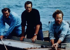 Three men stand on a small boat by blue water, one in a black shirt and two in light shirts, with a rope in the foreground.] - concise, but ensure under 125 chars.