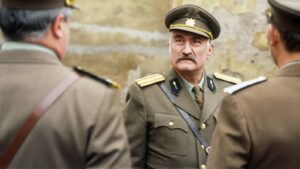 Older man in a vintage military uniform with a peaked cap and mustache speaks with two officers in tan uniforms outdoors.