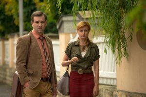 Two adults walk side by side on a street; the man wears a brown blazer and striped tie, the woman wears a green blouse and red plaid skirt with a belt, both carrying bags and looking ahead.