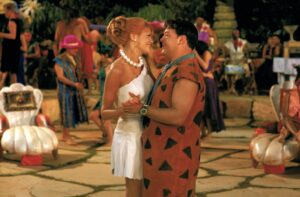 A bride in a white strapless dress with a pearl necklace dancing with a man in a brown spotted caveman costume at a lively costume party.