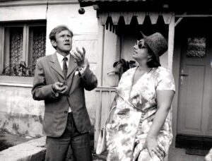Man in a suit gestures with his hands while speaking to a woman wearing a hat and sunglasses outside a house.