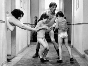 Adults and children in underwear playfully wrestling in a hallway, with a woman reaching in from the left.
