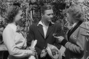 Three adults in vintage clothing converse outdoors; man in a suit holds a basket of flowers while two women chat nearby.