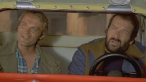 Two men in a vintage orange car, with the passenger smiling at the camera while the driver looks ahead.