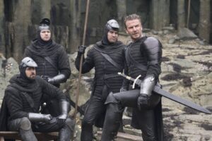 Four men in black medieval armor posing among rocky ruins; one kneels with a sword, three stand, all in dark protective gear.