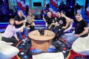 Group of seven people seated in a circle on a TV studio set, toasting with wine glasses; festive balloons and a band in the background.