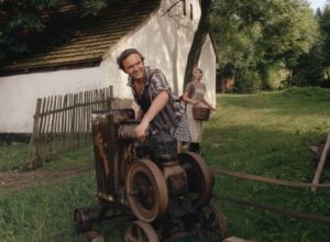 Man operating a rusty belt-driven machine on rail tracks in a rural yard, with a woman in an apron carrying a basket watching from behind a tree.