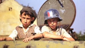 Two young boys lean over the edge of a weathered military tank, one wearing a cap and the other a helmet with a French flag sticker.