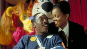 Smiling man in a blue outfit with face paint sharing a moment with a smiling woman in a black suit at a colorful party backdrop.