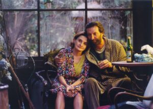Couple sitting close together by a window, the woman in a floral dress and the man in a checkered coat, holding glasses in a cozy room.