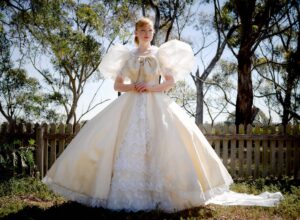 Woman in a vintage cream ballgown with lace underlay stands outdoors with clasped hands in front, beside a wooden picket fence.