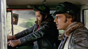 Two men ride in a pickup truck cab, one driving and the other in the passenger seat, both wearing leather jackets and caps.