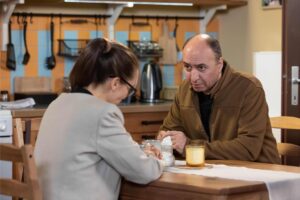 Two people sit across from each other at a wooden table, having a serious conversation in a cozy kitchen setting.