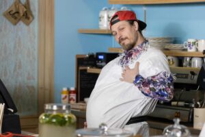 Smiling man in a silver poncho and colorful shirt leans toward the camera in a cafe.