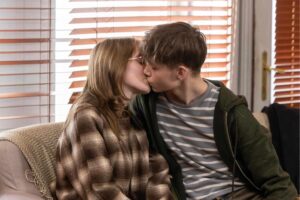 Couple kissing on a sofa indoors with horizontal wooden blinds behind them.