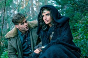 Man and woman sit in a dense forest; the woman wears a dark cloak and gold headpiece, looking ahead as if in a scene from a fantasy story.