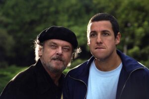 Two men posing outdoors in a park; the older man wears a black beret and has a gray beard, the younger man wears a blue jacket and light blue shirt.