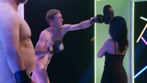 Shirtless man punches a pad held by a woman in a black dress in a neon-lit studio. Another shirtless man watches on the left.