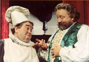 Two men in theatrical costumes share a playful moment on stage: one in a white chef’s outfit with a tall hat, the other in an ornate green vest, smiling at each other.