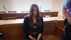 Young woman seated in a courtroom with hands restrained in front, wearing black, long brown hair and a serious expression.