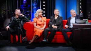 Four guests sit on a bright red couch on a TV talk-show set, smiling as they chat, with blue stage lights and a drummer in the background.