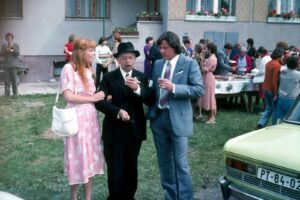 Group of three adults toasting at an outdoor party, with a pink-dressed woman on the left and two men in suits in the center, near a buffet table and a lime-green car in the foreground.