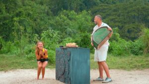 Young girl laughing in a yellow top crouches beside a man holding a large circular green shield outdoors.
