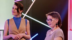 Two women on a brightly lit stage; left woman in a tan vest over blue top smiles, right woman with glasses talks. Neon lights in the background.