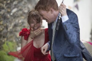 Smiling couple in formal attire dancing in the rain; woman in a red dress and man in a blue suit share a joyful moment.
