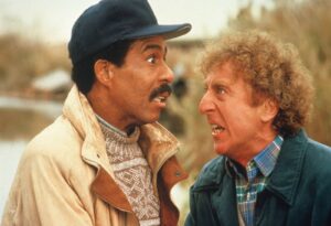 Two men argue closely outdoors; left man in beige jacket and cap, right man in plaid shirt and green jacket.