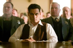 Centered man in white shirt and brown vest with a stern expression, seated at a table; two suited men with badges stand behind him.