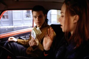 Two people in a car: a man counting a stack of cash while the woman gestures with her hand in the foreground, daytime street outside.