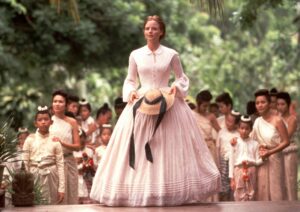 Woman in a pale pink dress rides a horse at the forefront of a parade, holding a hat with ribbons while children in white outfits walk behind in a lush garden setting.