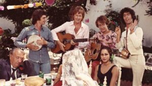 Outdoor garden party with musicians: two men strum guitars while a third holds a tambourine; decorations hang above. A veiled woman sits with her back turned at a table, and another woman in a black dress sits nearby, with an older man in a suit asleep at the table.