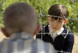 Boy with dark hair looks through barbed wire at a blurred person outdoors.