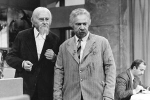 Two older men in suits stand together indoors; one with a white beard smiles, the other looks surprised in a striped-gray blazer with stains.