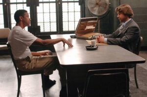Interrogation scene: man in a white T-shirt and khakis sits across from a suited man at a metal table, with an open briefcase between them.