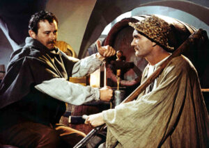 Two men in historical robes shake hands across a wooden workbench in a dim workshop, with a large grinding wheel behind them.