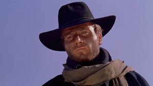 Man in a wide-brim black hat and beige scarf against a clear blue sky, looking straight ahead.