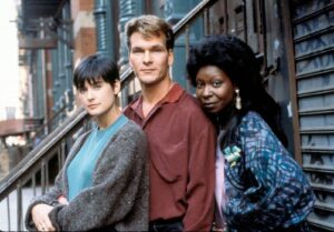 Three friends pose together on an urban stoop, woman left in teal top and gray cardigan, man center in red shirt, woman right with large hair and patterned jacket.