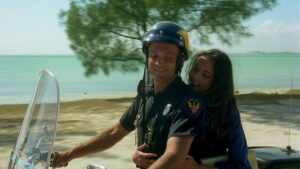 Police officer wearing a helmet rides a motorcycle on a beach, with a smiling woman embracing him from behind.