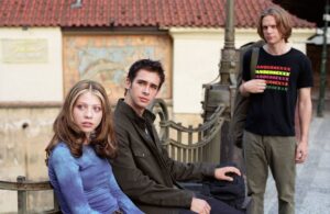 Two young adults sit on a bench while a third person stands nearby on a sunny street with a red-tiled roof in the background.