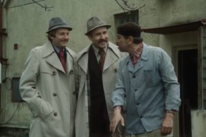 Three men in trench coats and hats stand outside a weathered building, chatting and smiling at each other.